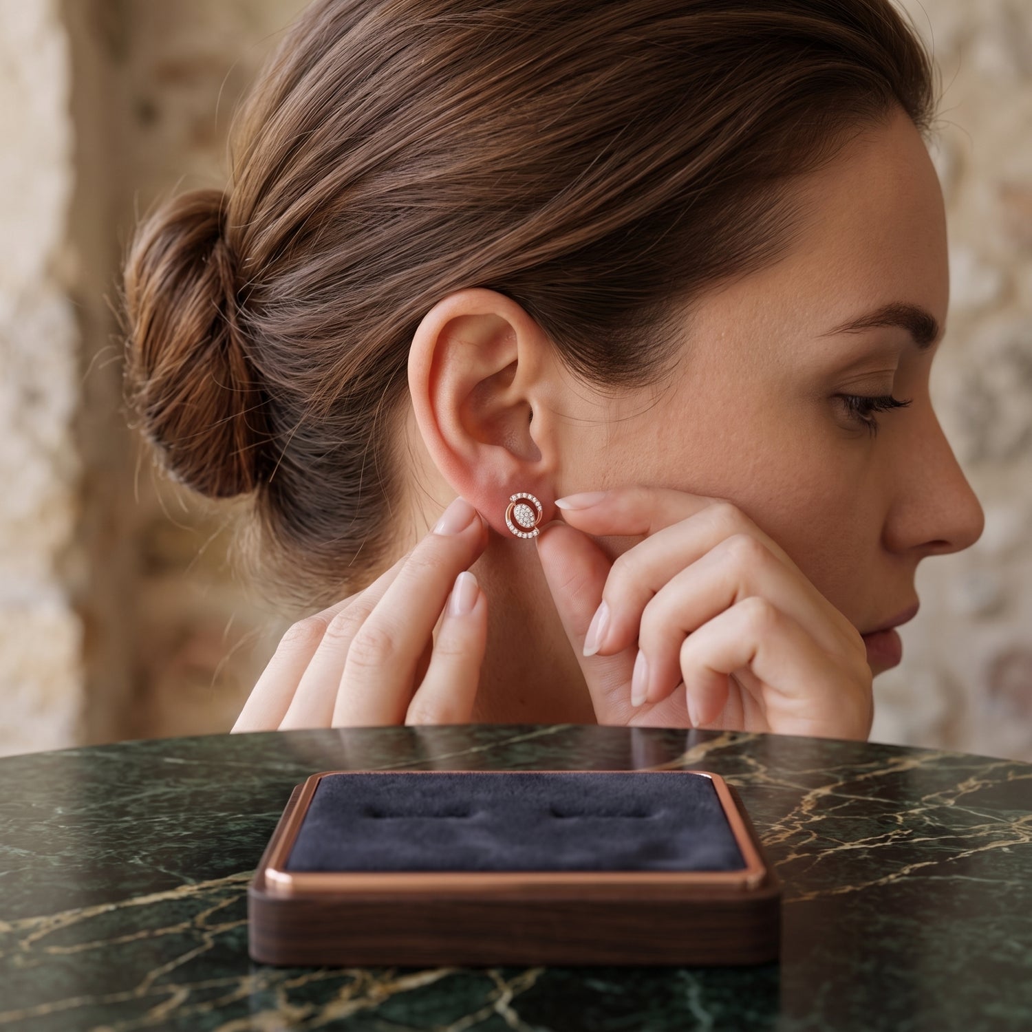 A woman in profile wearing a small round stud earring with a white center and a rose-gold-toned halo, with her fingers touching the earring. A dark velvet jewelry box sits on a green marble table in the foreground.