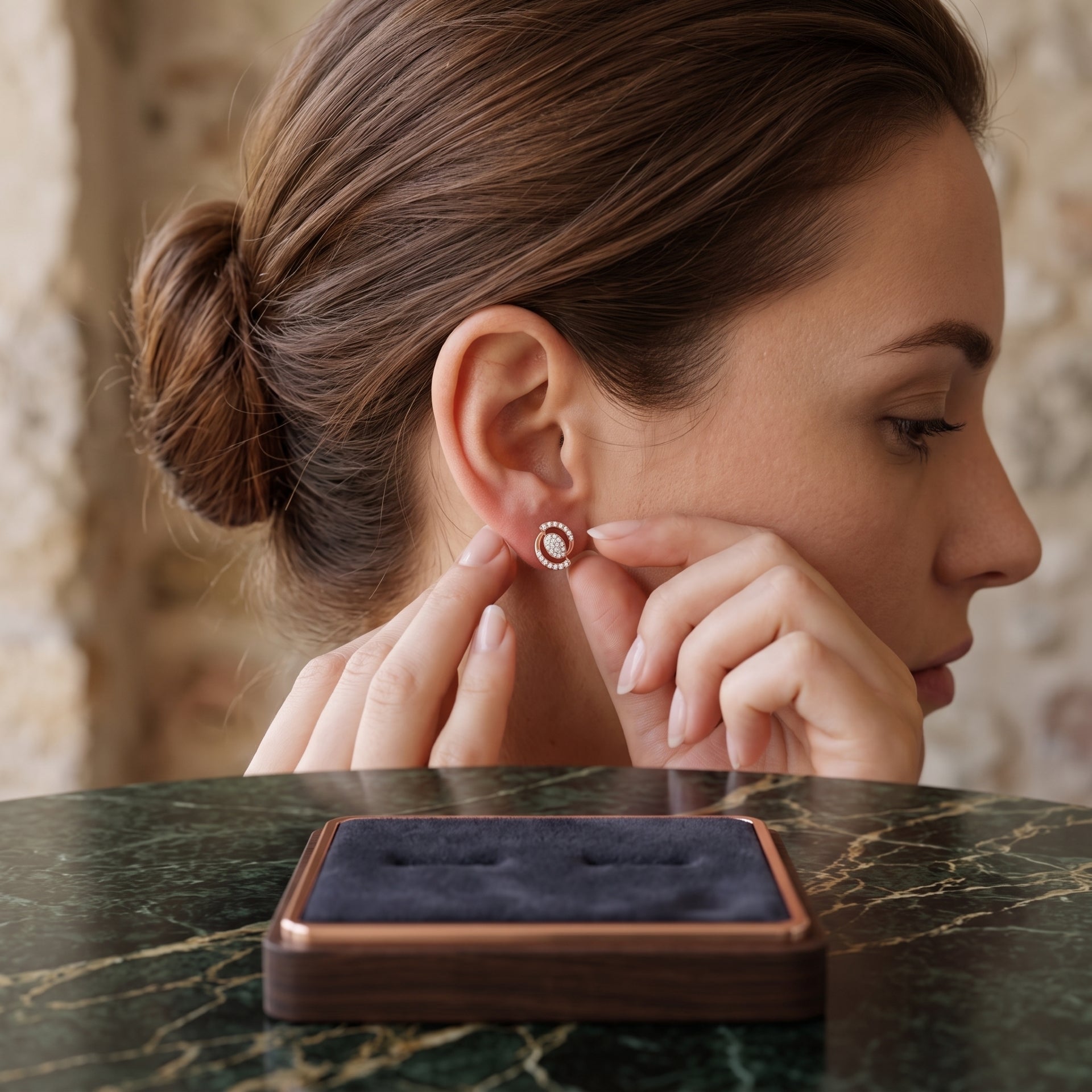 A woman in profile wearing a small round stud earring with a white center and a rose-gold-toned halo, with her fingers touching the earring. A dark velvet jewelry box sits on a green marble table in the foreground.