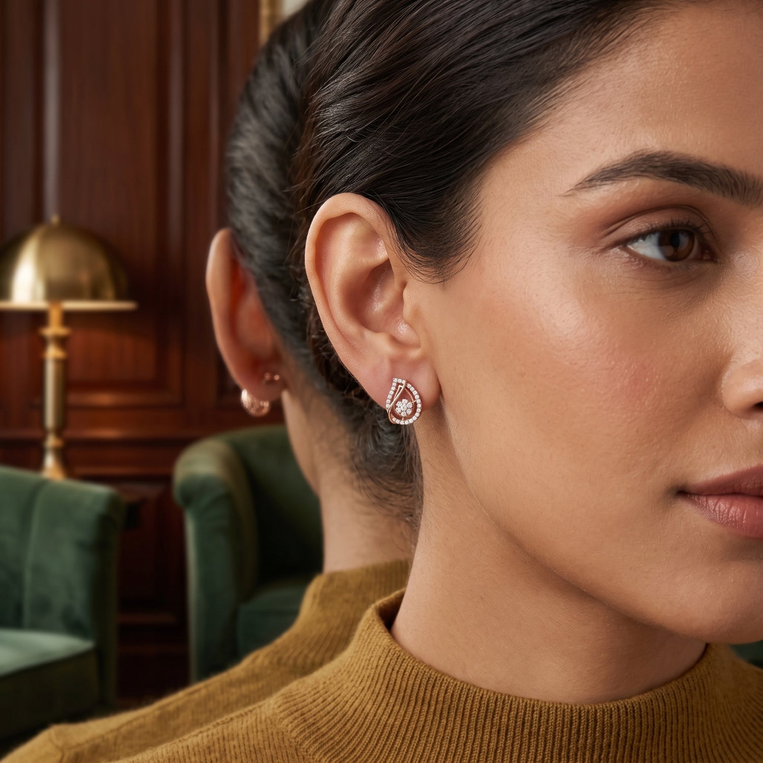 A close-up lifestyle shot of a woman wearing a teardrop-shaped silver stud earring with a clear central gemstone and a halo of small pink stones. The background shows a warm, dark wood-paneled room with a green armchair.