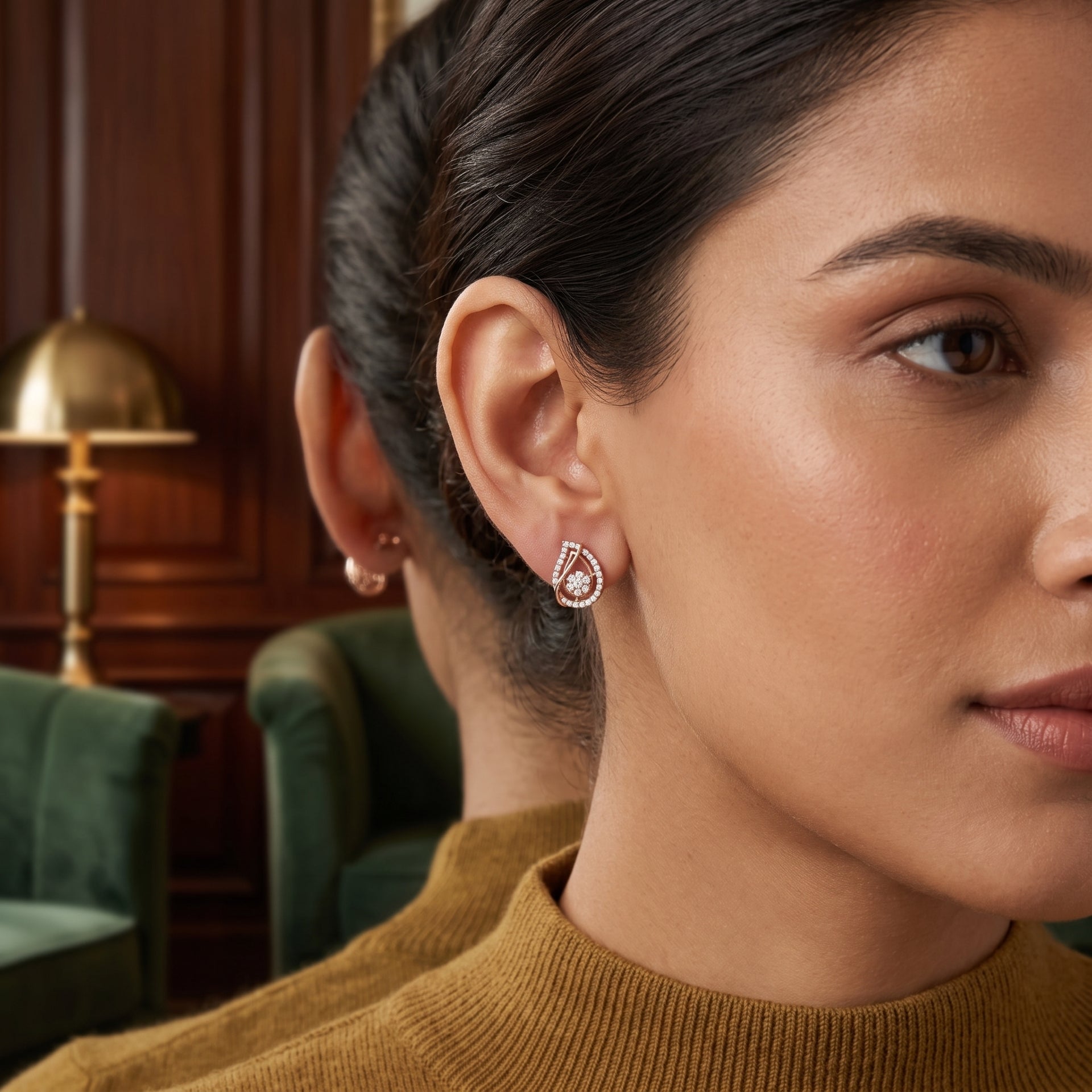 A close-up lifestyle shot of a woman wearing a teardrop-shaped silver stud earring with a clear central gemstone and a halo of small pink stones. The background shows a warm, dark wood-paneled room with a green armchair.