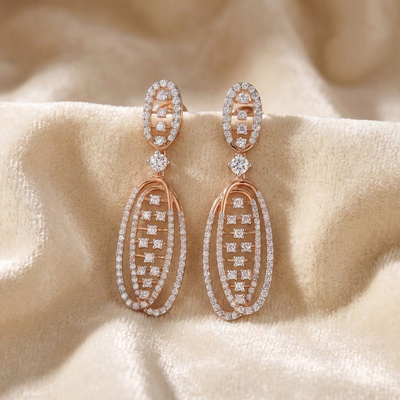 Rose gold drop earrings with multiple rows of clear pavé-set stones, displayed on a soft beige satin fabric background.