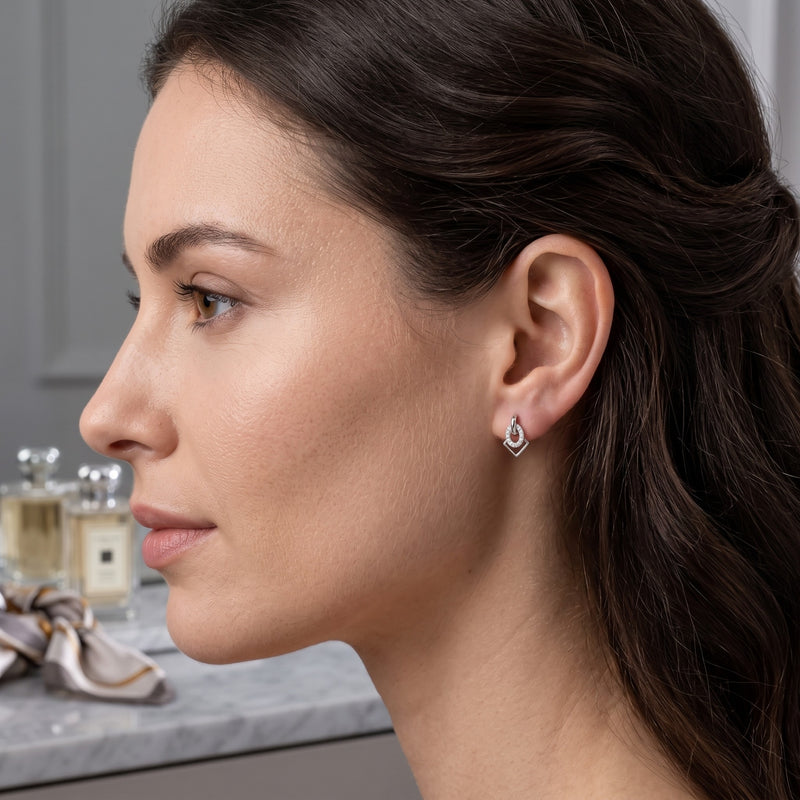 A close-up side profile of a woman wearing a small silver teardrop stud earring with clear gemstones, set against a softly blurred bathroom background with perfume bottles.
