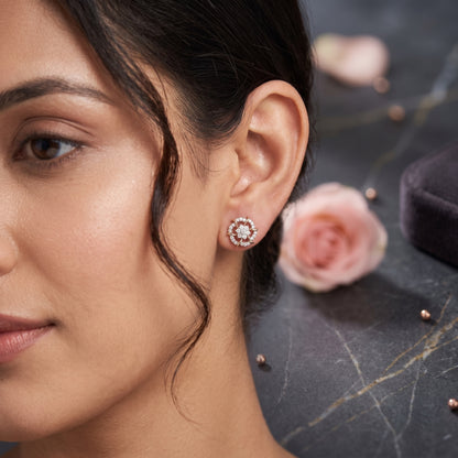 A close-up lifestyle shot of a woman wearing a rose gold stud earring with a halo of small clear stones, set against a dark marble surface with soft pink rose petals.