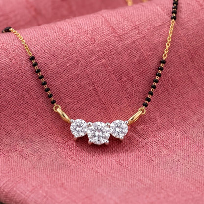 A close-up of a delicate necklace draped on pink textured fabric. The necklace features a gold chain with small black beads and a central cluster of three clear round-cut gemstones in a gold setting.