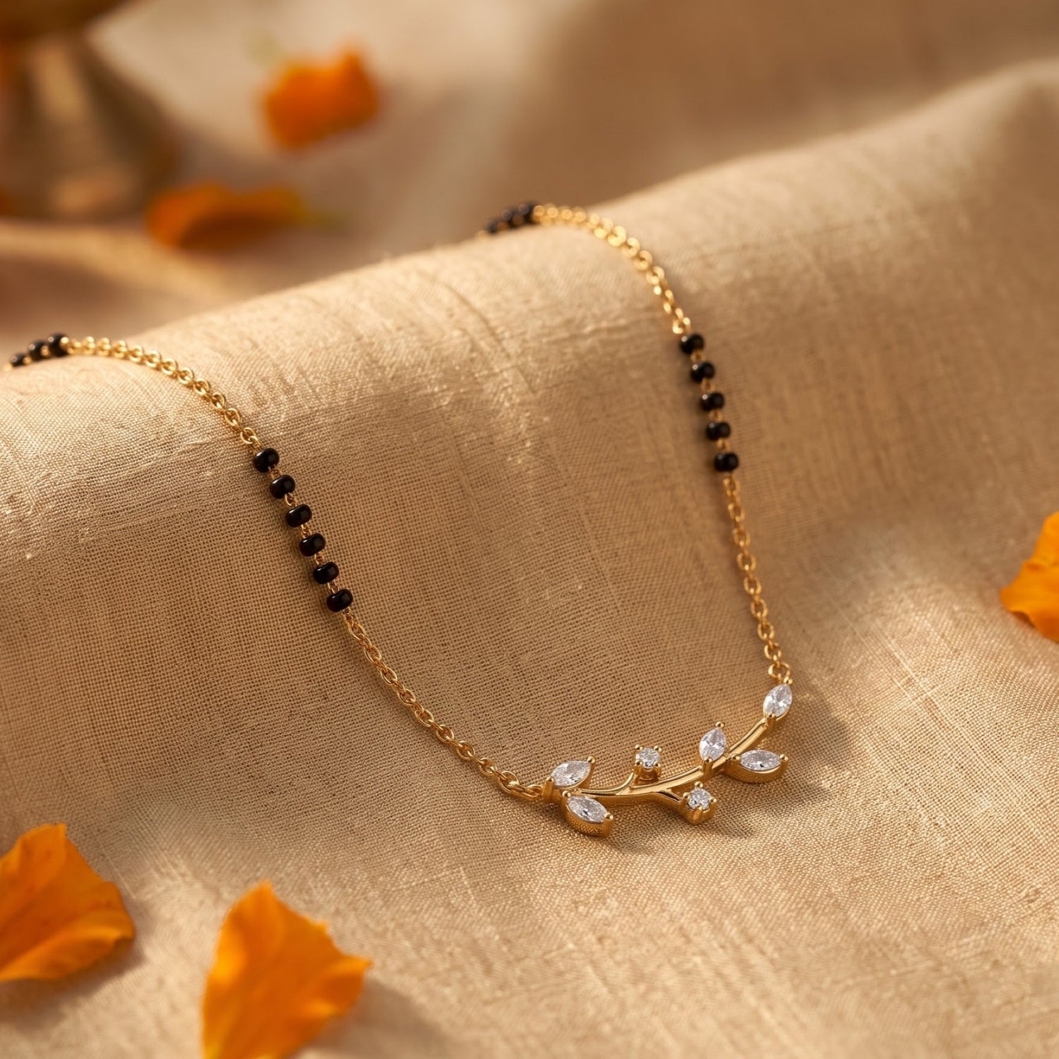 A delicate gold chain necklace with small black beads and a leaf-shaped pendant set with clear stones, draped on beige textured fabric with scattered orange petals.