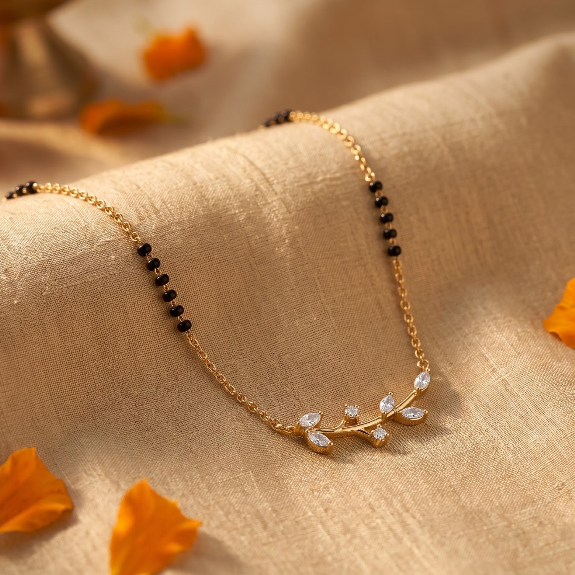 A delicate gold chain necklace with small black beads and a leaf-shaped pendant set with clear stones, draped on beige textured fabric with scattered orange petals.