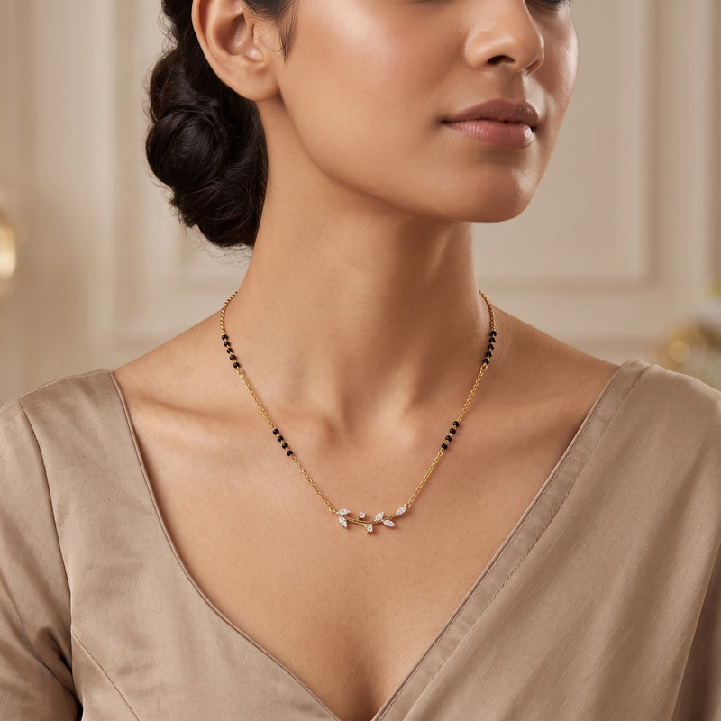 A close-up of a woman wearing a delicate gold necklace with small black beads and a central white floral or leaf-like cluster pendant, paired with a taupe satin top.