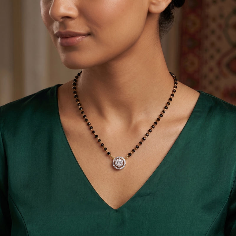 A close-up of a woman wearing a beaded necklace with a round silver-toned pendant featuring a central crystal or diamond, paired with a dark green satin V-neck top.