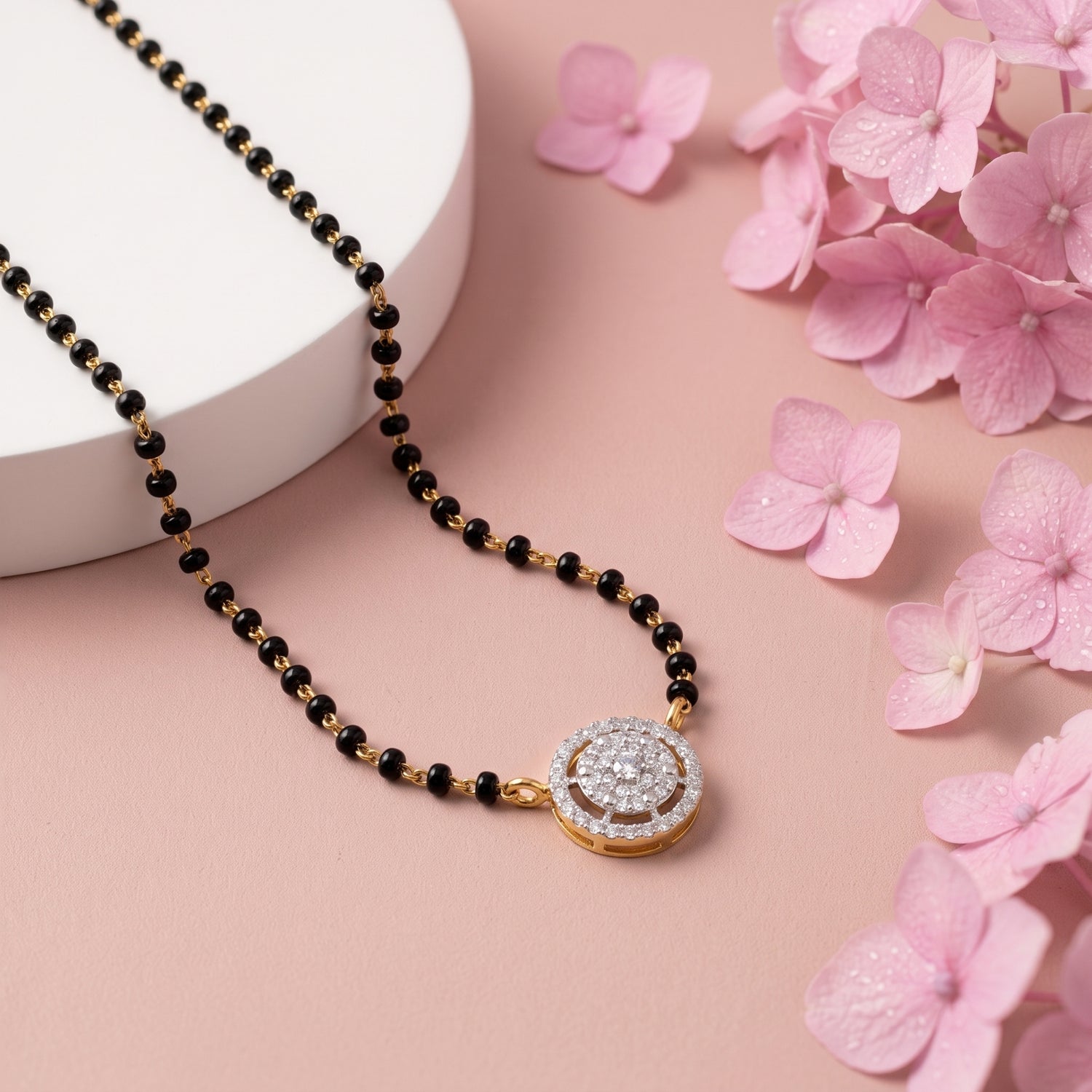 A gold and black beaded necklace with a round pavé-set pendant, styled on a pink background with a white cylindrical prop and pink flowers.