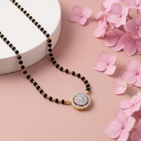 A gold and black beaded necklace with a round pavé-set pendant, styled on a pink background with a white cylindrical prop and pink flowers.