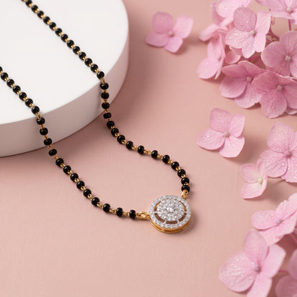 A gold and black beaded necklace with a round pavé-set pendant, styled on a pink background with a white cylindrical prop and pink flowers.
