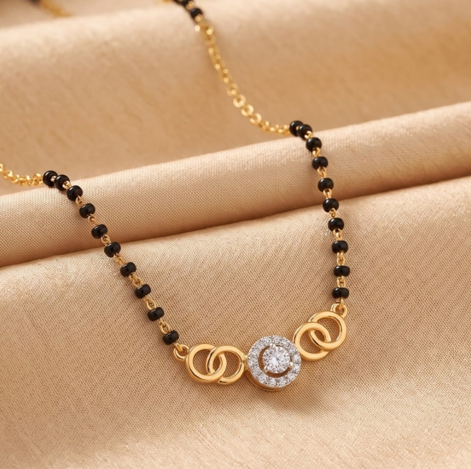 A gold and black beaded necklace with a central circular pendant featuring a clear stone, displayed on a beige satin fabric background.