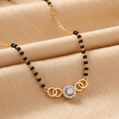 A gold and black beaded necklace with a central circular pendant featuring a clear stone, displayed on a beige satin fabric background.