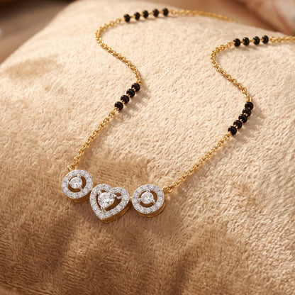 A gold chain necklace with a beaded section of small black beads and a pendant featuring three heart and circle motifs paved with clear stones, displayed on a textured beige fabric surface.