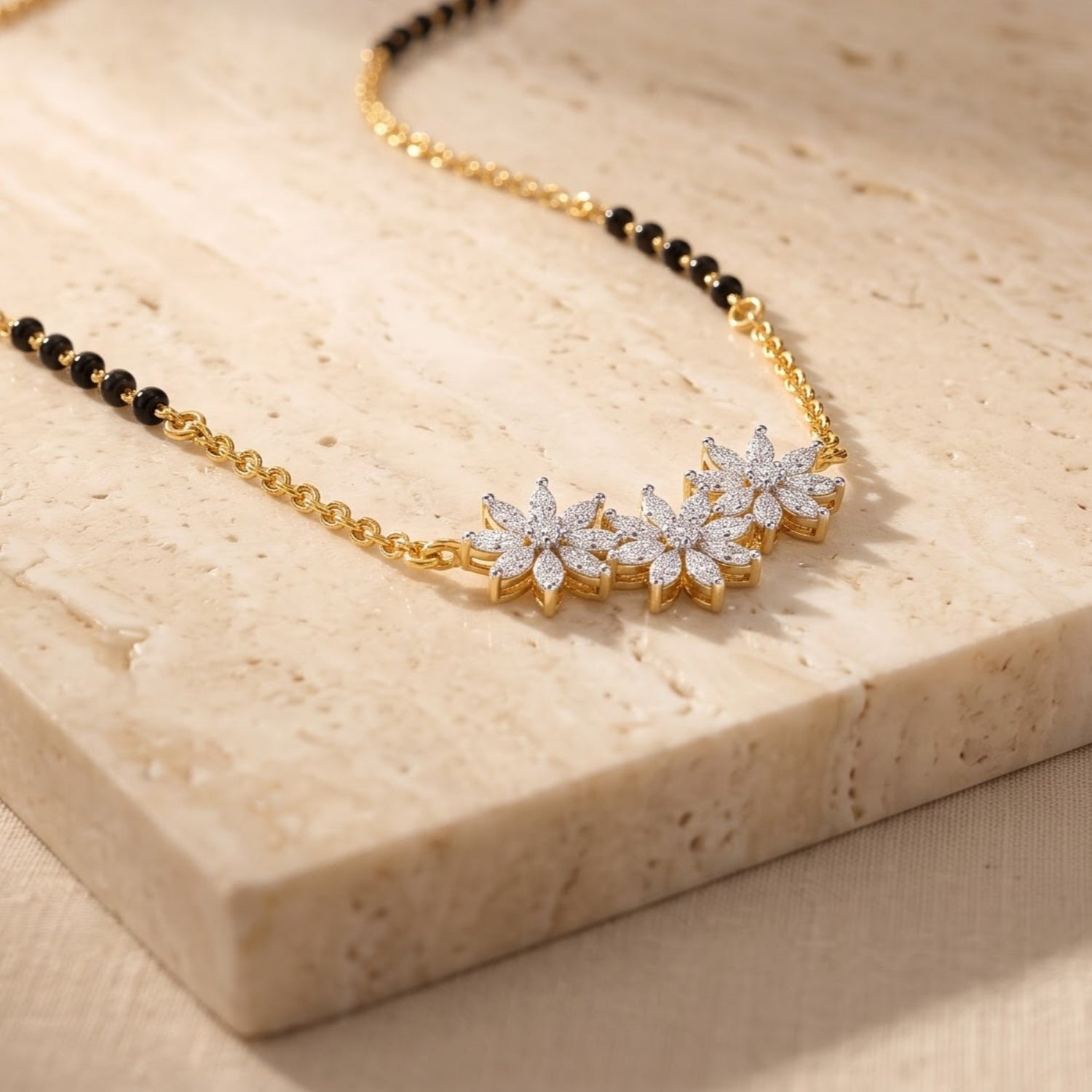 A gold chain necklace with a row of three flower-shaped clusters set with clear stones, displayed on a beige stone slab.