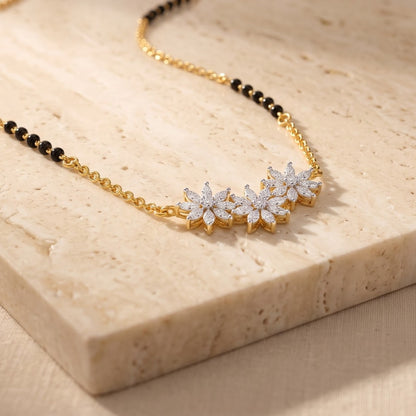 A gold chain necklace with a row of three flower-shaped clusters set with clear stones, displayed on a beige stone slab.