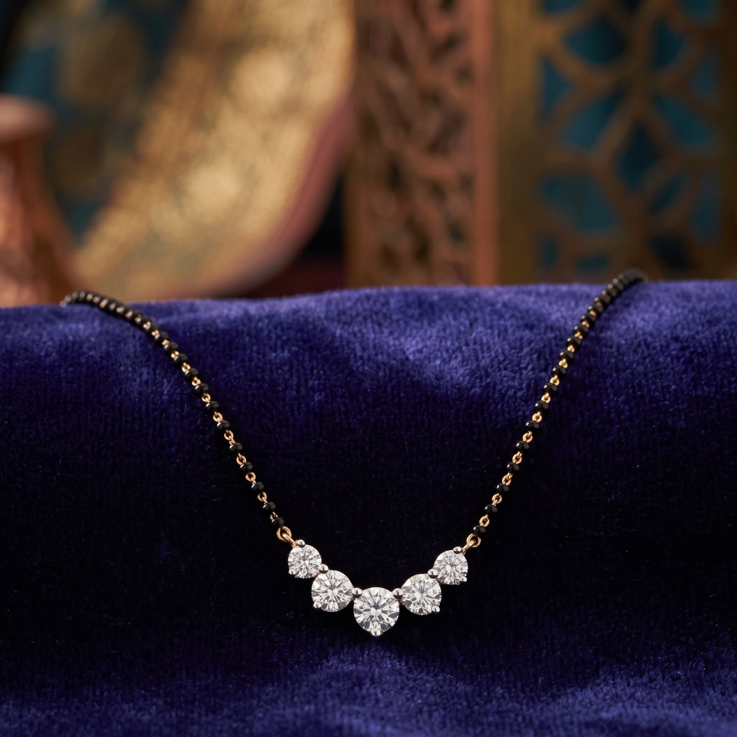 A delicate necklace with a small cluster of clear stones on a fine gold-toned chain, displayed on a deep blue velvet surface with a softly blurred ornate background.