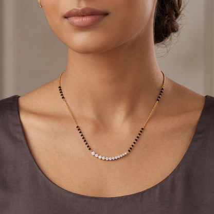 A close-up of a model wearing a delicate necklace with a curved row of small white stones set on a gold chain, accented with small black beads.
