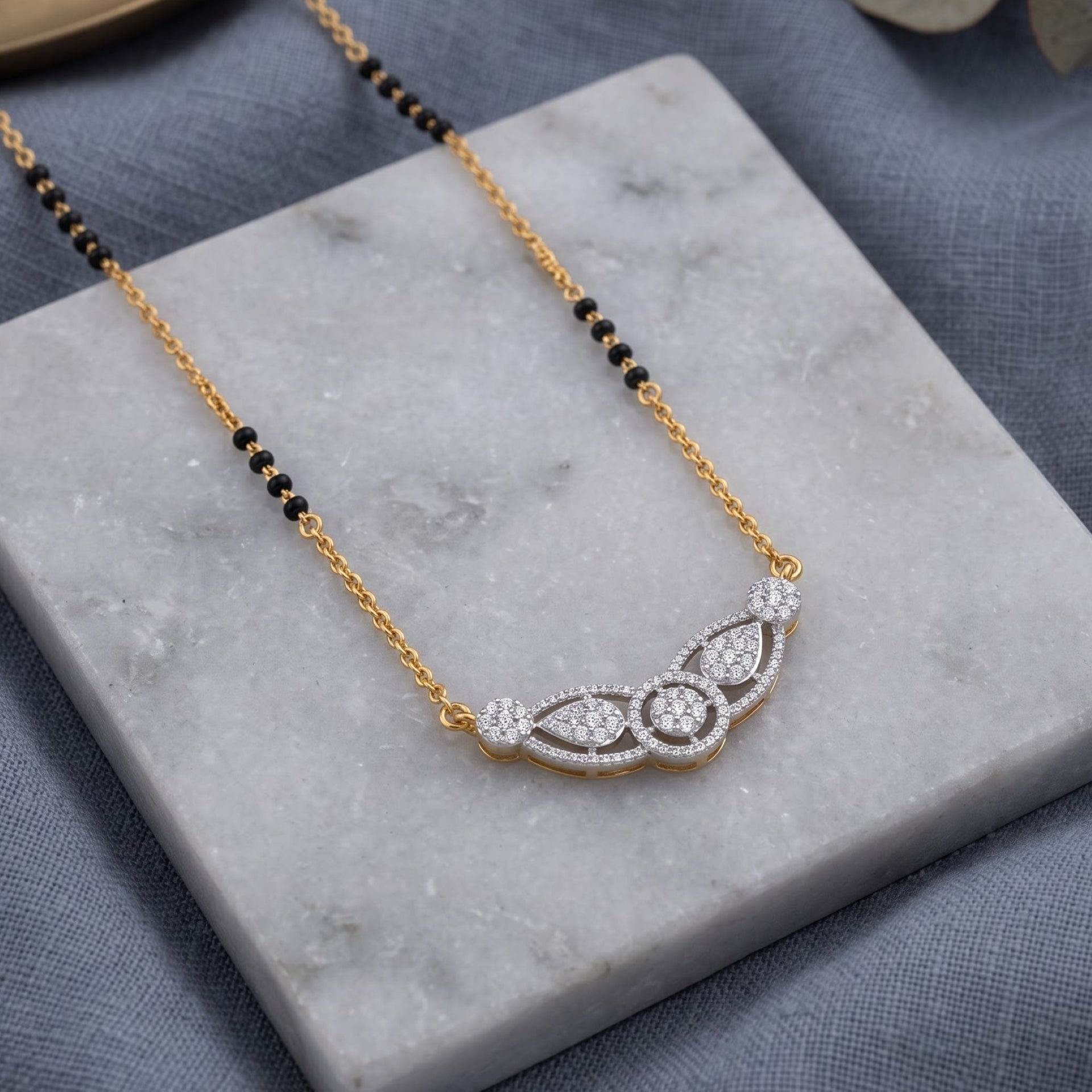 A gold and white gold necklace with a beaded black chain and a curved pavé pendant, displayed on a white marble block over a textured gray fabric background.