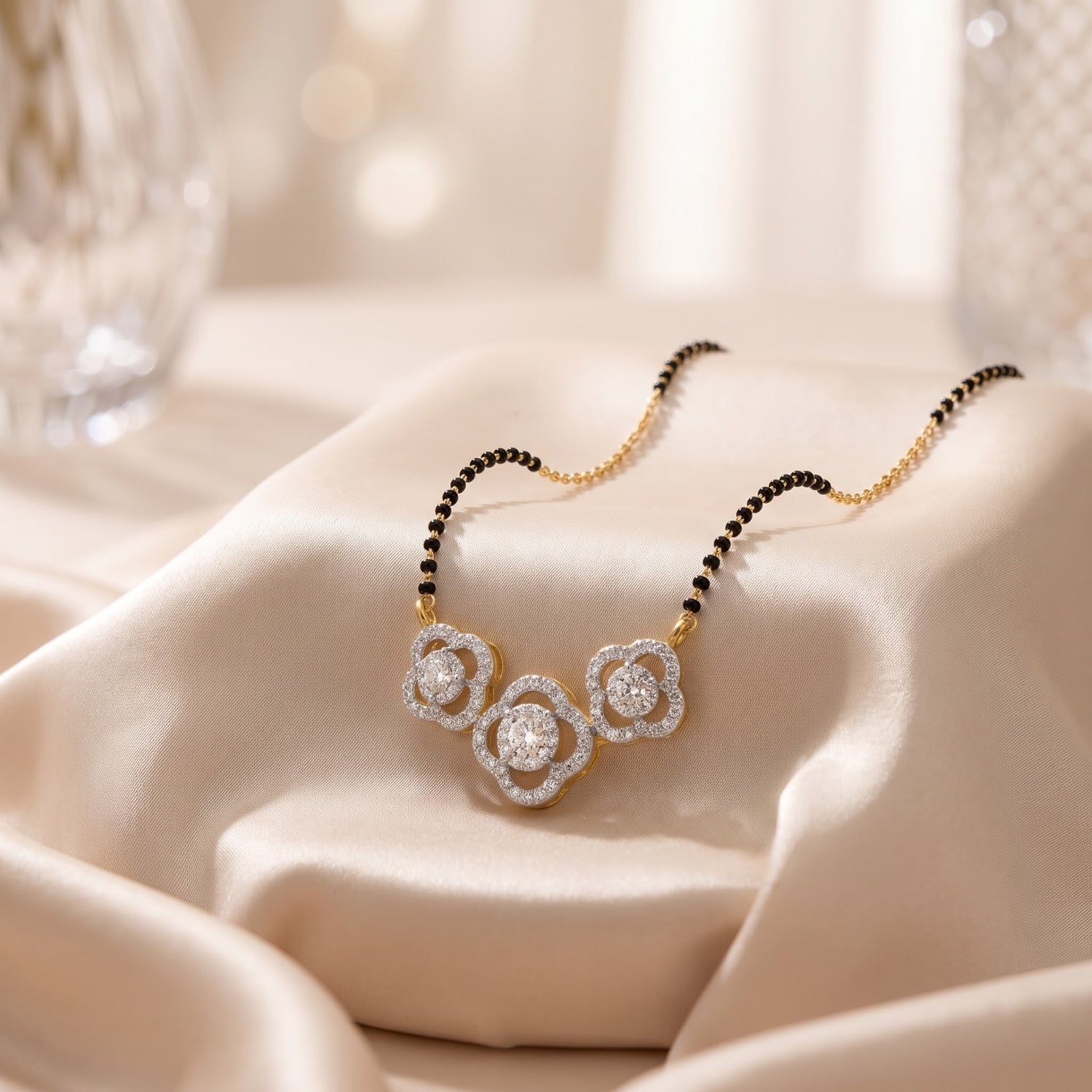 A close-up of a delicate necklace draped on soft cream satin fabric. The necklace has a gold-toned chain with small black beads on one side and a three-flower pendant set with clear stones.