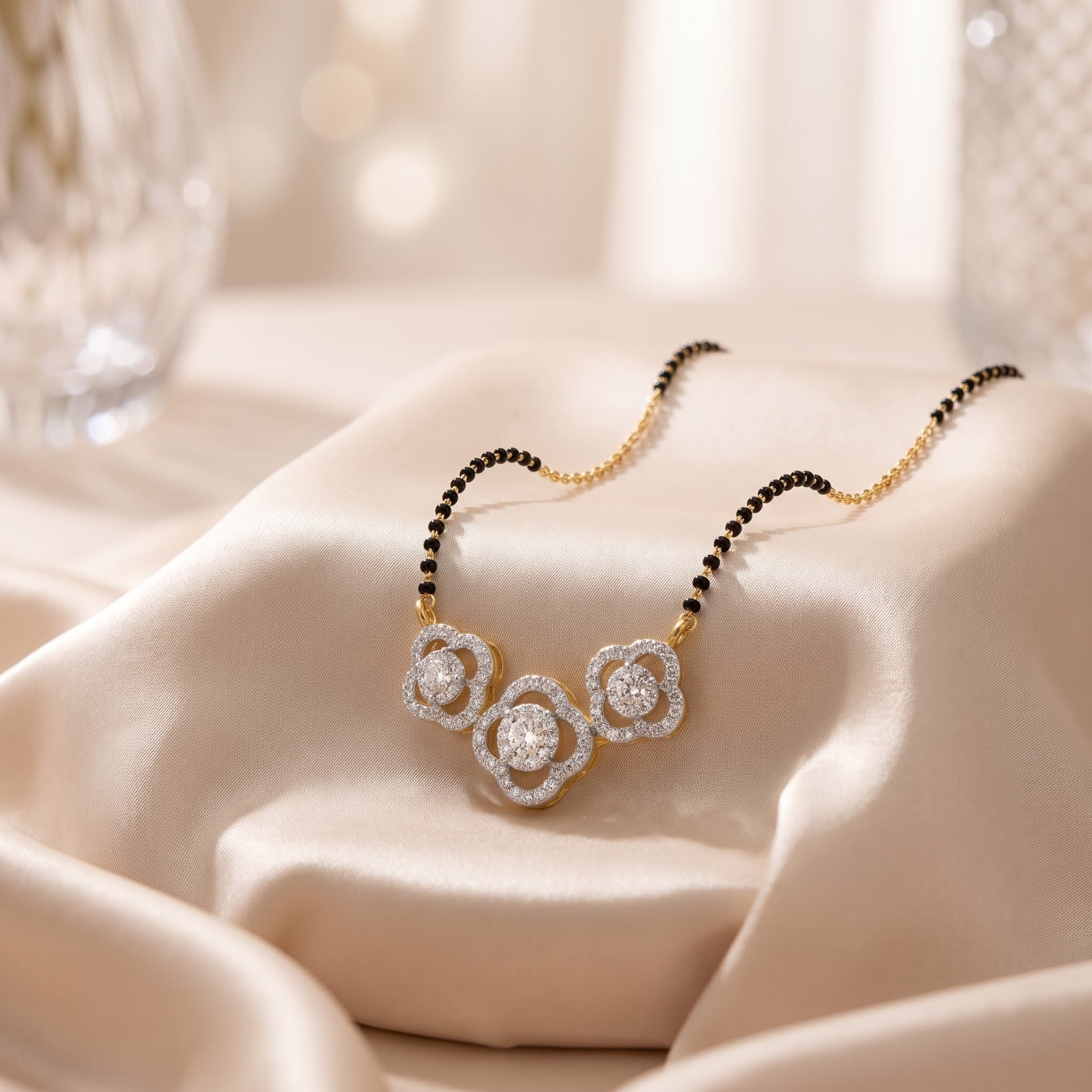 A close-up of a delicate necklace draped on soft cream satin fabric. The necklace has a gold-toned chain with small black beads on one side and a three-flower pendant set with clear stones.