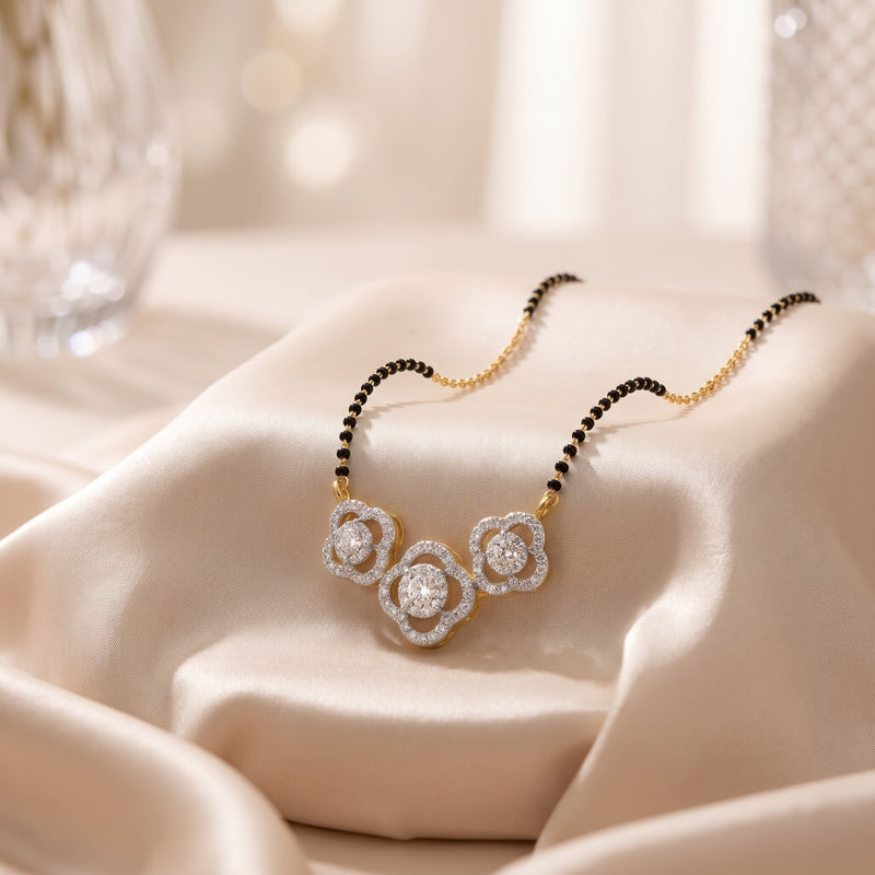 A close-up of a delicate necklace draped on soft cream satin fabric. The necklace has a gold-toned chain with small black beads on one side and a three-flower pendant set with clear stones.
