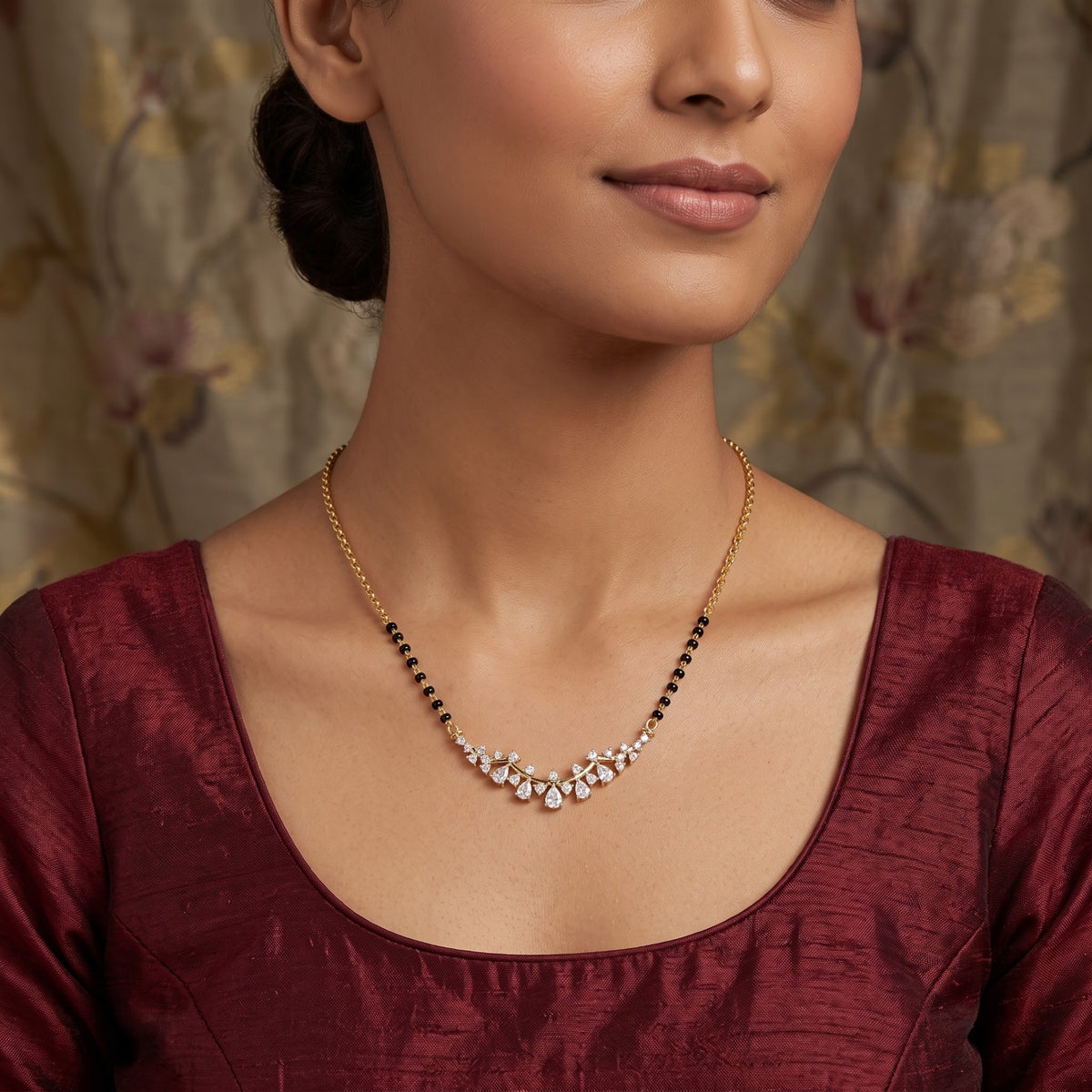 A close-up of a woman wearing a delicate gold necklace with a curved cluster of clear stones and small black beads along the chain, paired with a deep maroon blouse.