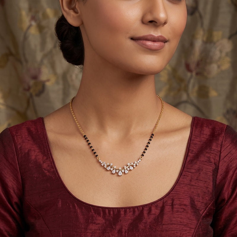 A close-up of a woman wearing a delicate gold necklace with a curved cluster of clear stones and small black beads along the chain, paired with a deep maroon blouse.