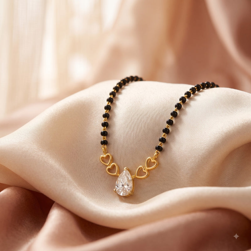 A delicate necklace with a black and gold beaded chain, featuring three gold heart outlines and a teardrop-shaped clear gemstone pendant, displayed on draped satin fabric.