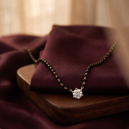 A delicate gold chain necklace with a round clear gemstone pendant, draped elegantly on a dark burgundy fabric resting on a wooden tray.
