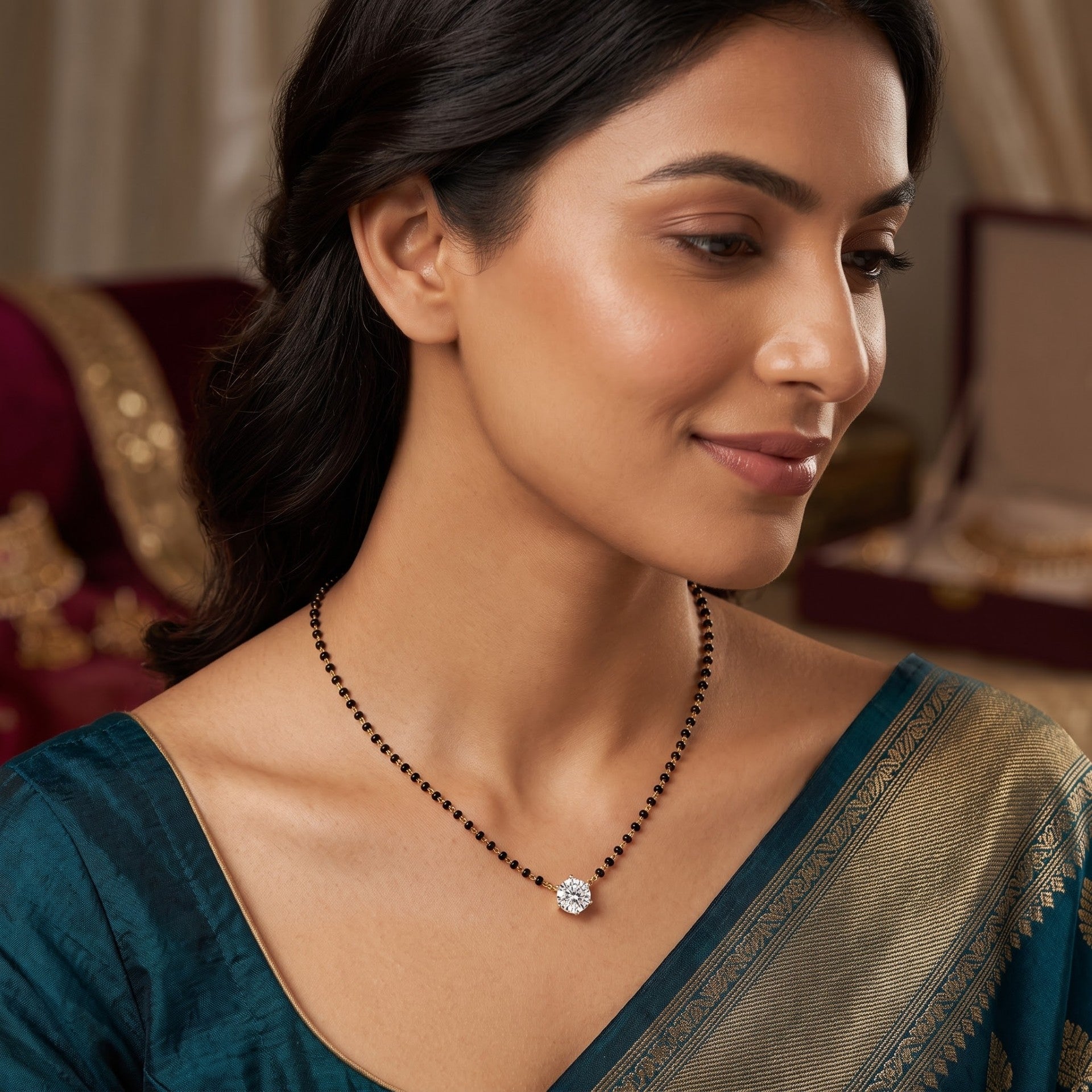 A woman wearing a black beaded necklace with a small white stone pendant, styled with a teal and gold saree, shown in a warm, elegant indoor setting.