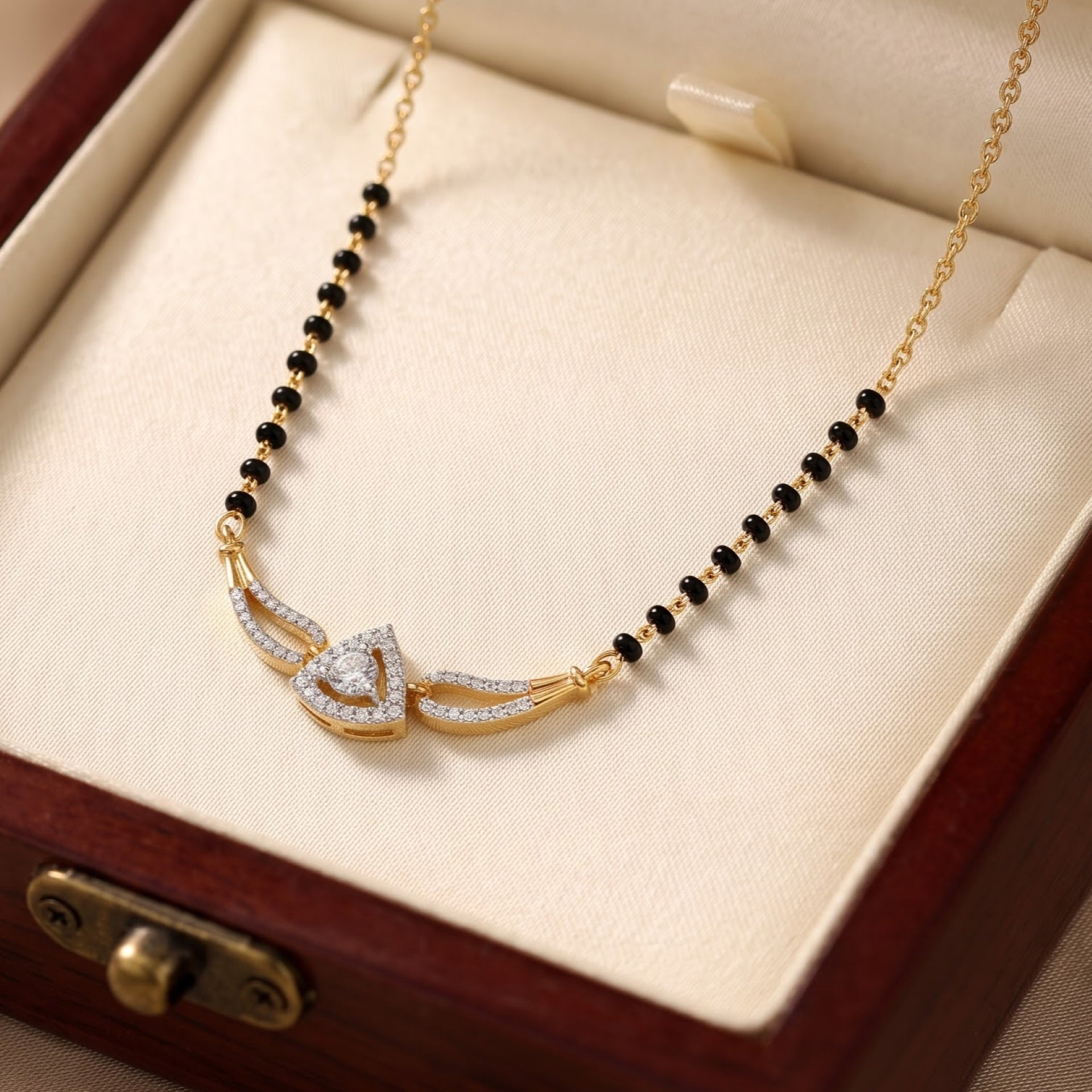 Gold chain necklace with a central gold pendant featuring a heart-shaped crystal design, flanked by curved crystal-accented wings, displayed on a cream jewelry box insert.