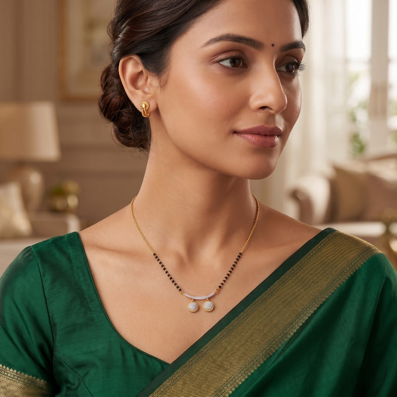 A woman wearing a gold necklace with a silver crescent and two white pearls, paired with a green saree, in a softly lit indoor setting.