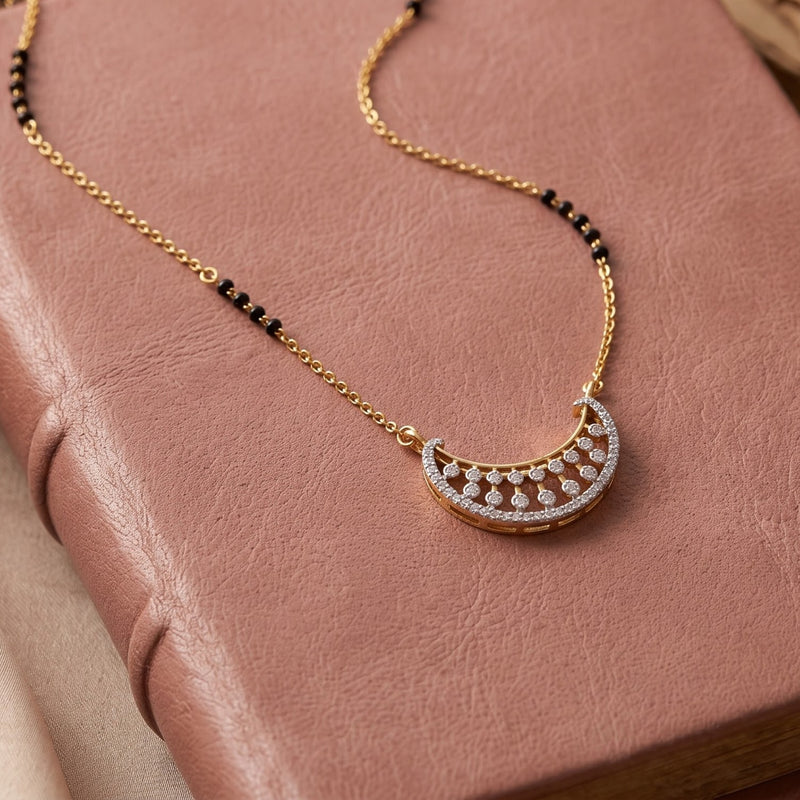 A gold crescent moon pendant necklace with pavé-set clear stones and a beaded chain with black and gold beads, displayed on a textured dusty pink leather surface.