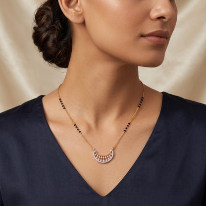 A close-up of a woman wearing a delicate gold necklace with a crescent-shaped pendant featuring small pink and white stones, paired with a navy blue top against a soft beige satin background.