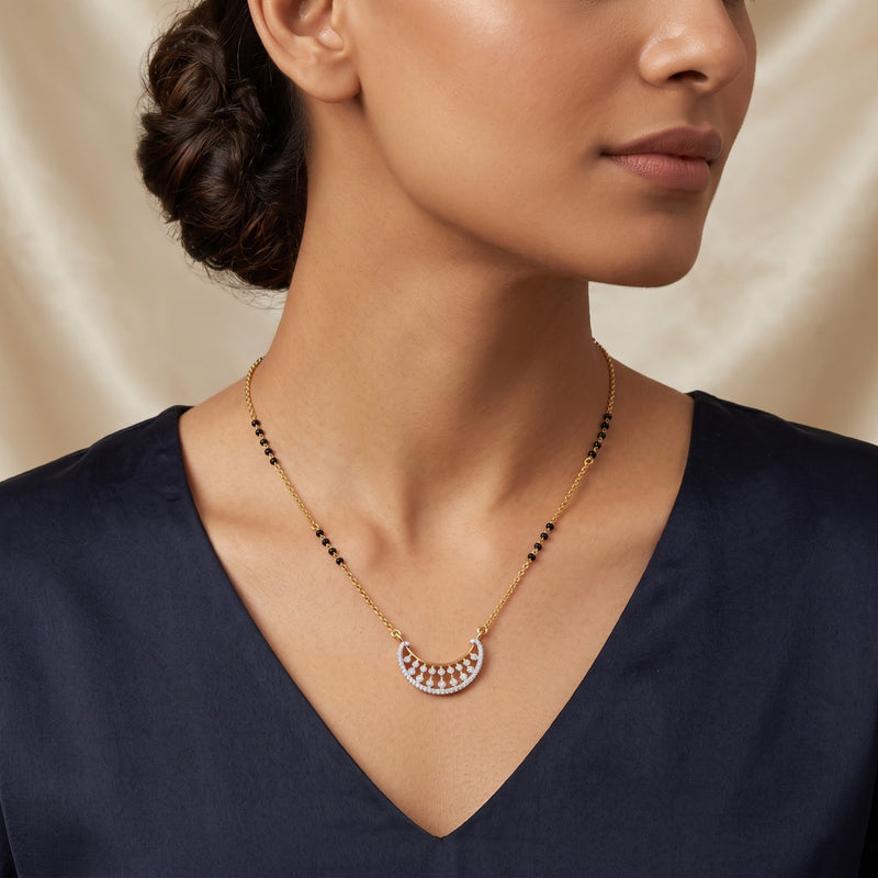 A close-up of a woman wearing a delicate gold necklace with a crescent-shaped pendant featuring small pink and white stones, paired with a navy blue top against a soft beige satin background.