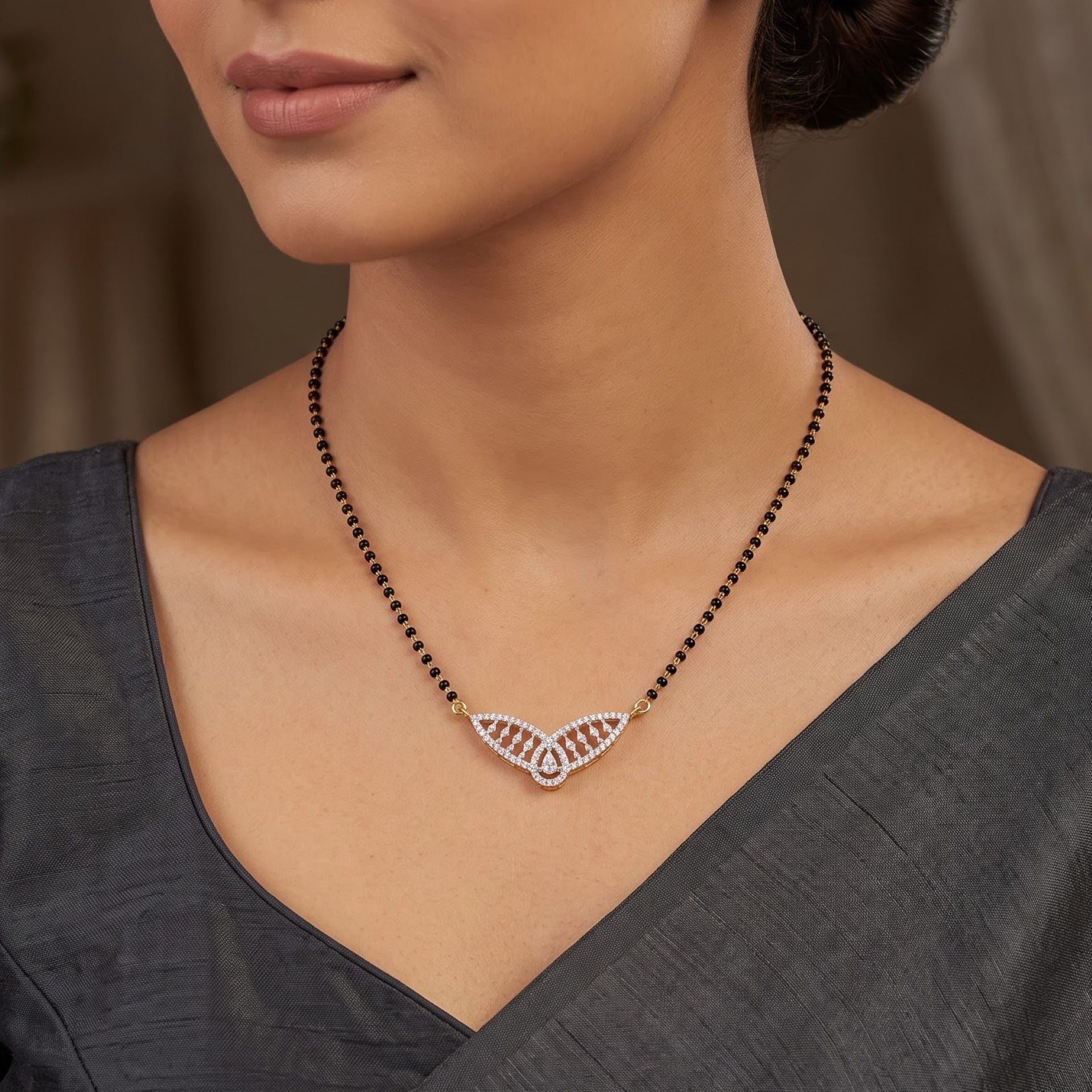 A close-up of a woman wearing a black beaded necklace with a wing-shaped pendant set with small clear stones and red stones, paired with a dark gray V-neck top.