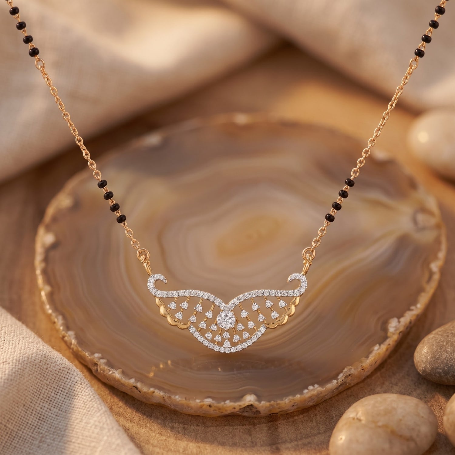 Gold necklace with a wing-like pendant set with small clear stones, displayed on a polished agate slice with beige fabric and stones in the background.