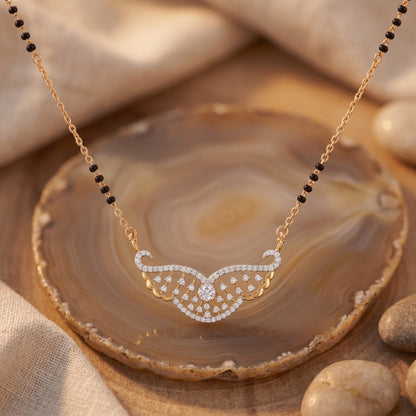 Gold necklace with a wing-like pendant set with small clear stones, displayed on a polished agate slice with beige fabric and stones in the background.