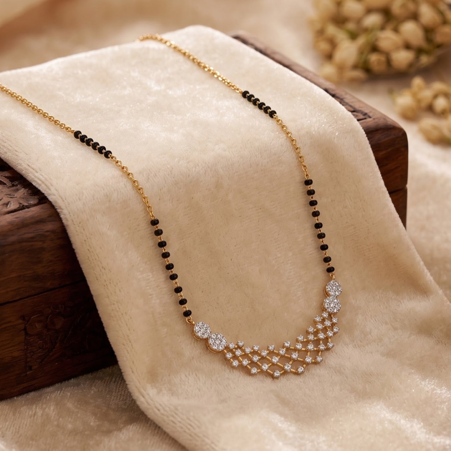 A gold chain necklace with black bead accents and a sparkling crystal geometric pendant, draped on a beige velvet fabric over a dark wooden block.