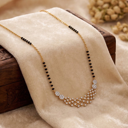 A gold chain necklace with black bead accents and a sparkling crystal geometric pendant, draped on a beige velvet fabric over a dark wooden block.