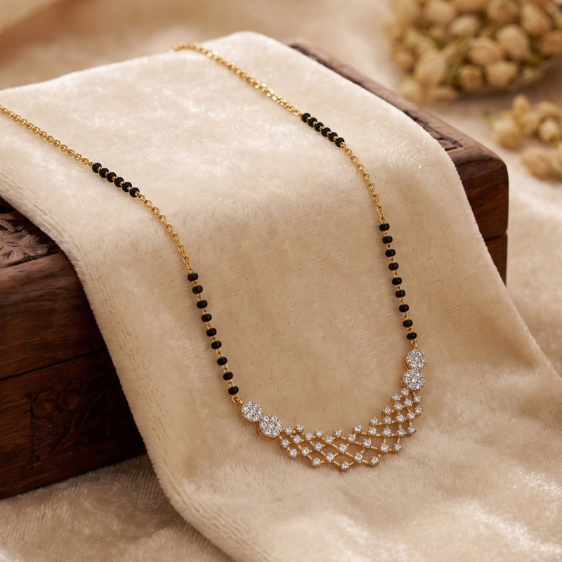A gold chain necklace with black bead accents and a sparkling crystal geometric pendant, draped on a beige velvet fabric over a dark wooden block.