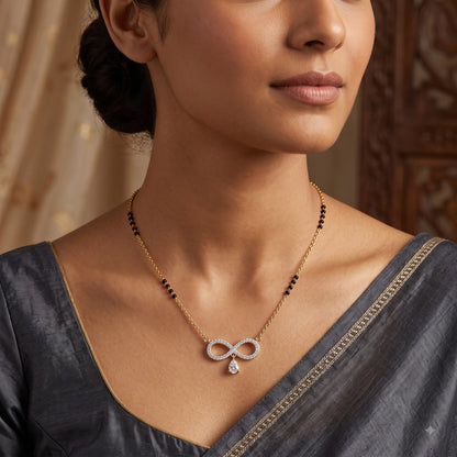 A close-up lifestyle shot of a woman wearing a delicate gold chain necklace with a silver infinity symbol pendant set with small clear stones, and a small teardrop crystal drop. The necklace has black bead accents on the chain.