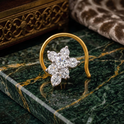 A gold crescent-shaped nose ring with a flower cluster of clear gemstones, displayed on a green marble surface.