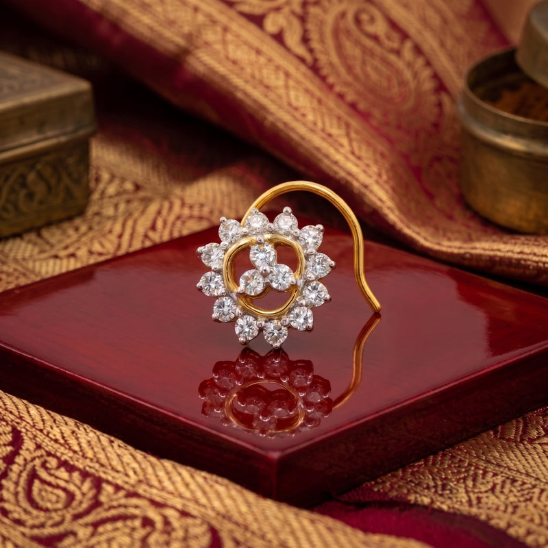A gold and diamond nose ring displayed on a glossy dark red square tray, surrounded by ornate gold and red patterned fabric.