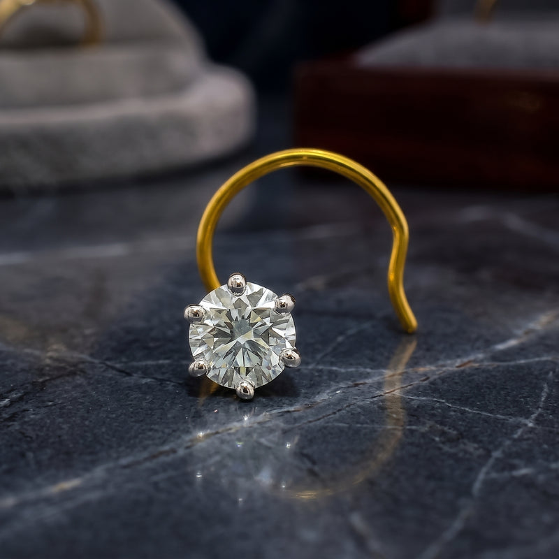 A single round-cut diamond solitaire nosepin with a yellow gold prong setting and a curved gold band, resting on a dark marble surface.