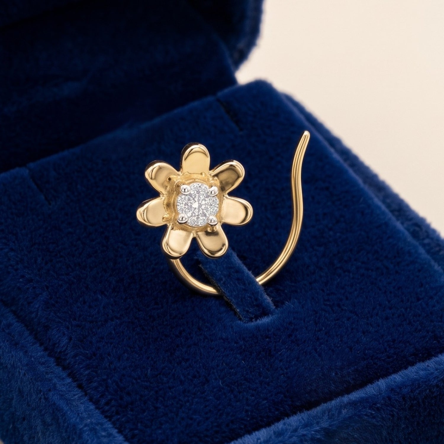 A gold flower-shaped nose ring with a central cluster of clear gemstones, displayed on a deep blue velvet ring box.