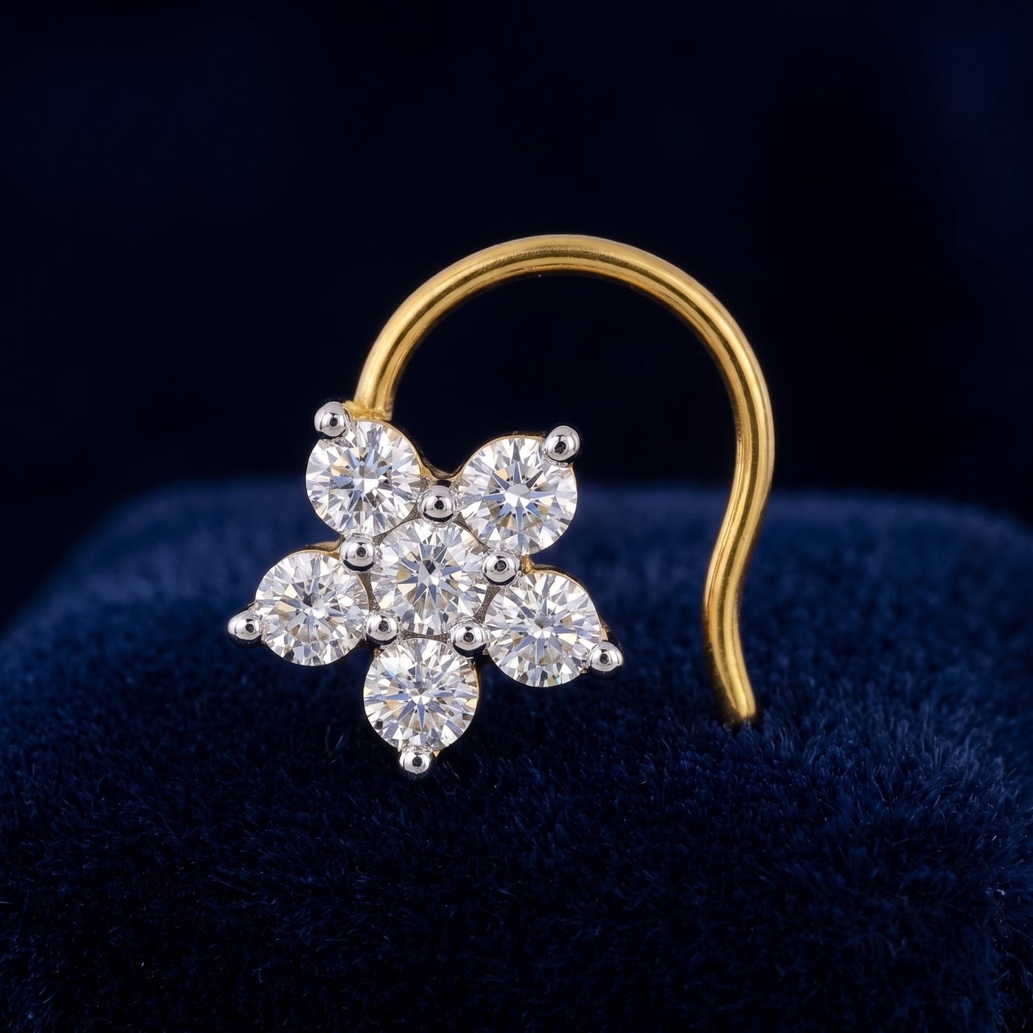 A gold nose ring featuring a flower-shaped cluster of clear round-cut gemstones, displayed on a dark blue velvet surface.