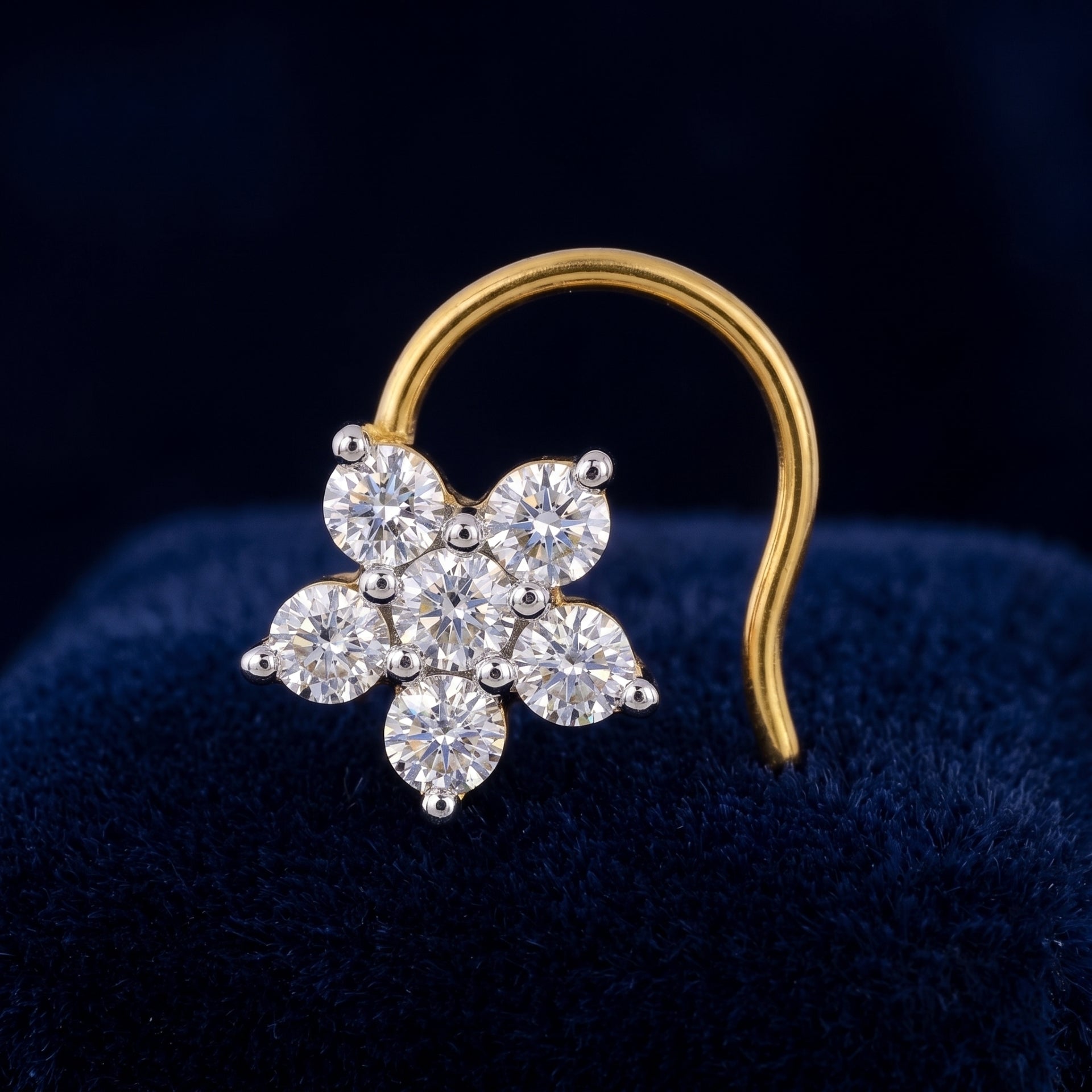 A gold nose ring featuring a flower-shaped cluster of clear round-cut gemstones, displayed on a dark blue velvet surface.