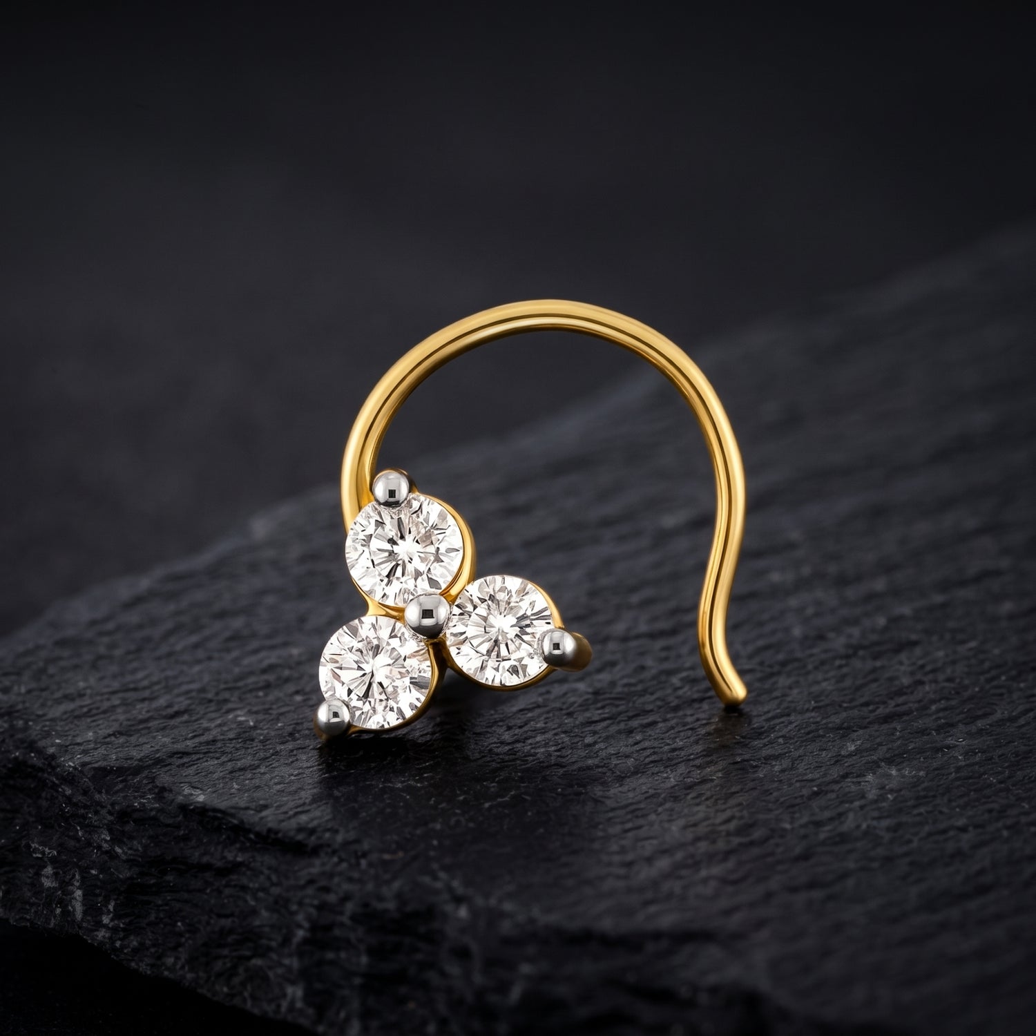 A gold crescent-shaped nose ring with three round clear gemstones, displayed on a dark textured slate surface.